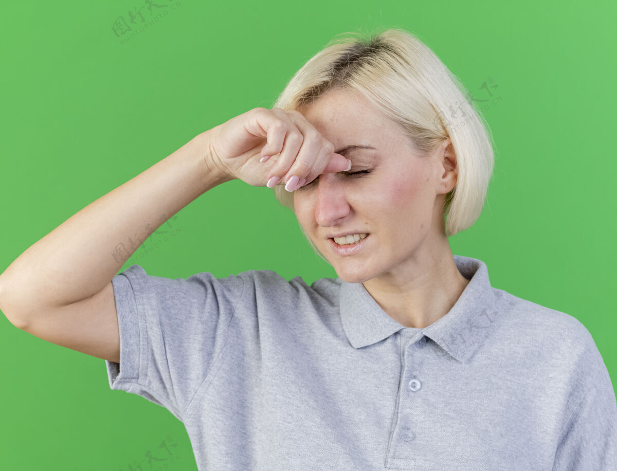 姿势疼痛的年轻金发病女人闭着眼睛站着 把手放在隔离在绿色墙上的额头上疾病人女人