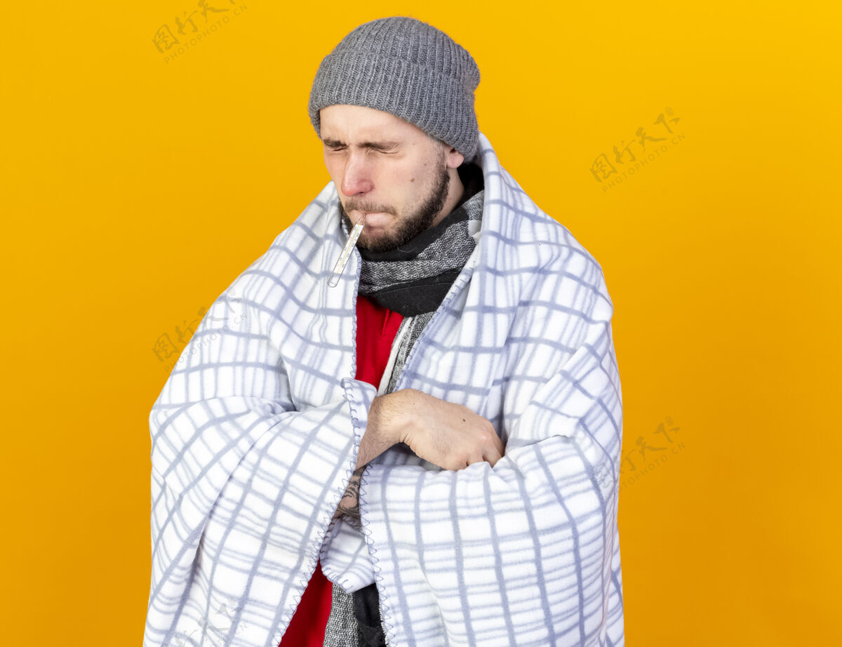 交叉一个戴着冬日帽 戴着围巾 交叉着双臂 站在格子布架子上的年轻病人把体温计放在嘴里 隔离在橙色的墙上年轻人人围巾