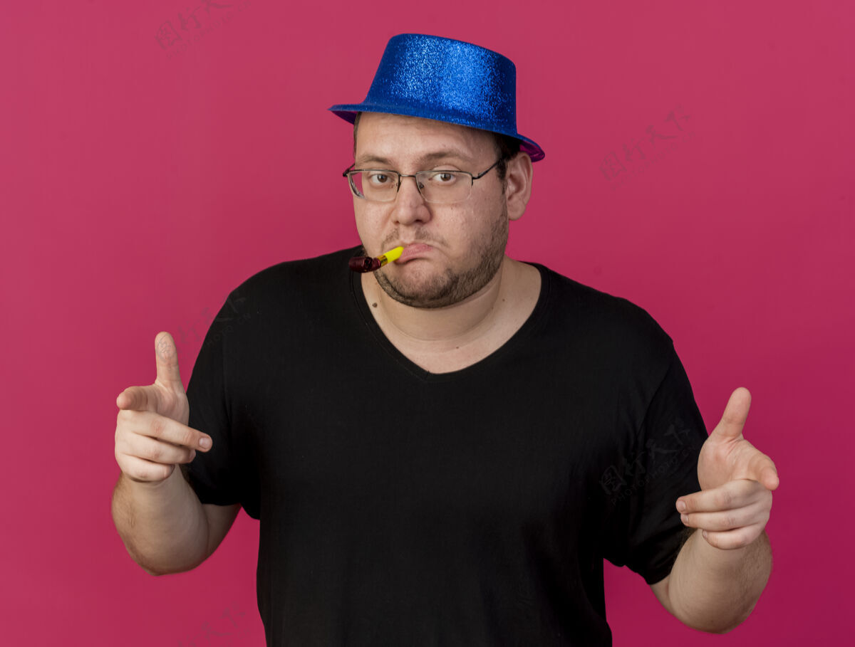 人戴着眼镜 戴着蓝色派对帽的自信的成年男子指着前面吹着派对哨子 隔离在粉红色的墙上光学公民穿