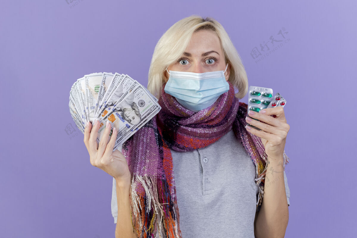 穿着震惊的年轻金发病妇女戴着医疗面具和围巾持有金钱和医疗药包隔离在紫色的墙壁市民医疗女人