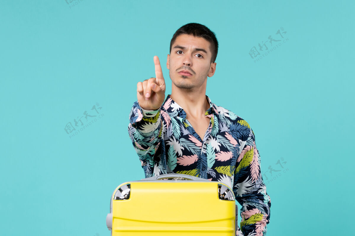 展示年轻的男性正准备度假 黄色的袋子在蓝色的墙上露出他的手指假期旅行航行