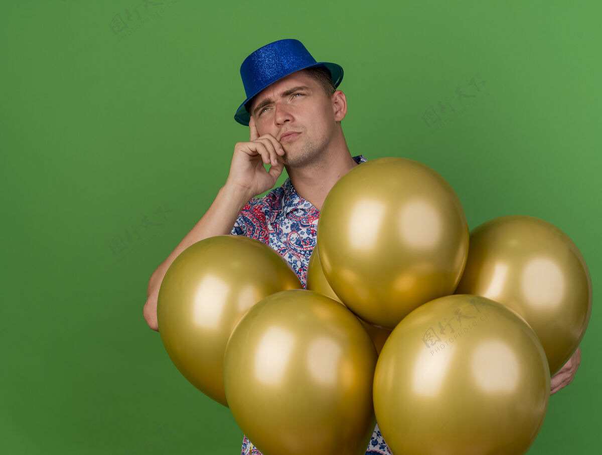 困惑困惑的年轻人戴着蓝色帽子站在气球后面 把手放在绿色的脸颊上穿帽子年轻
