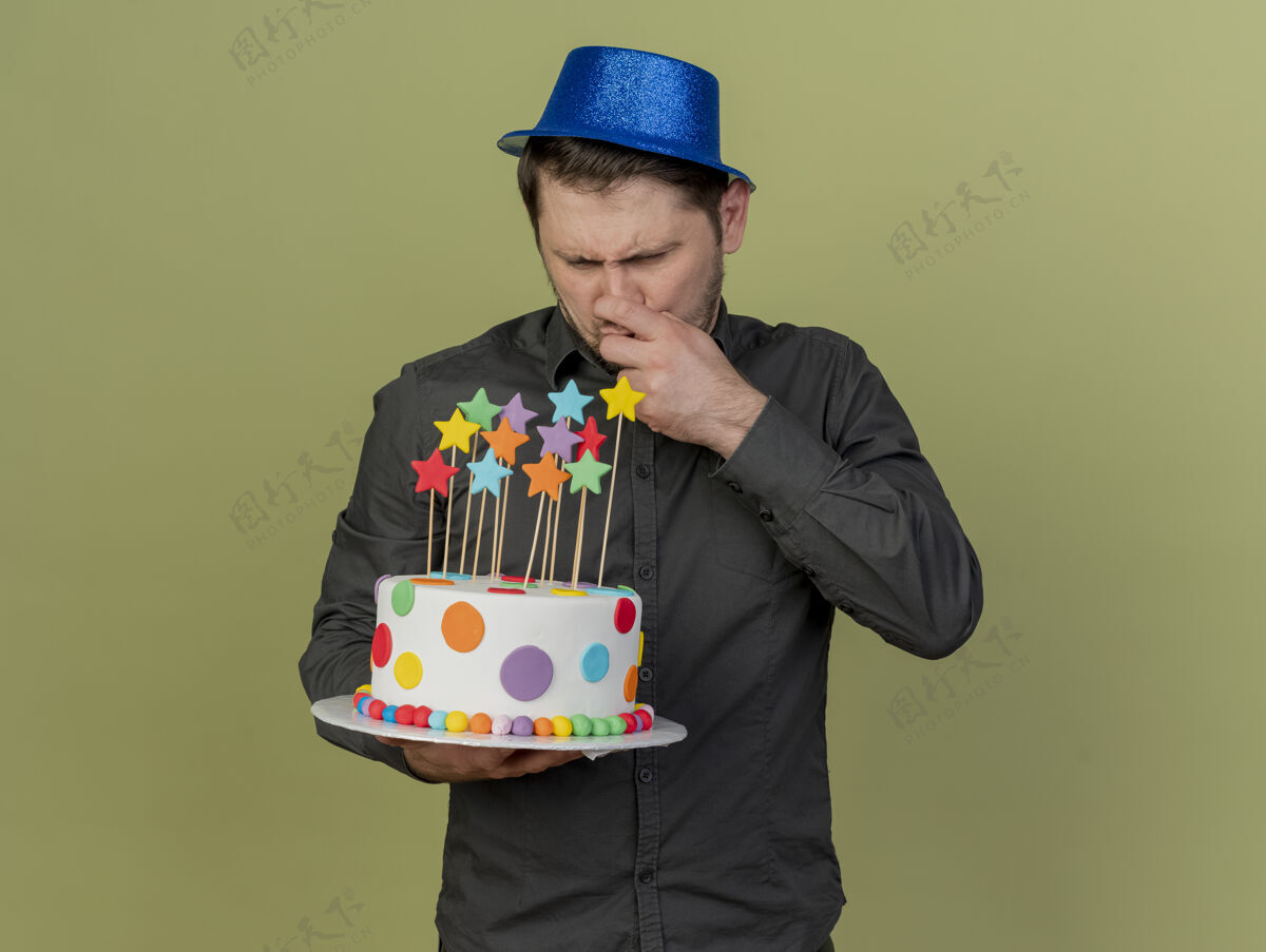 绿色一个体贴的年轻人 穿着黑色衬衫 戴着蓝色帽子 拿着蛋糕看着橄榄绿的鼻子年轻家伙思考