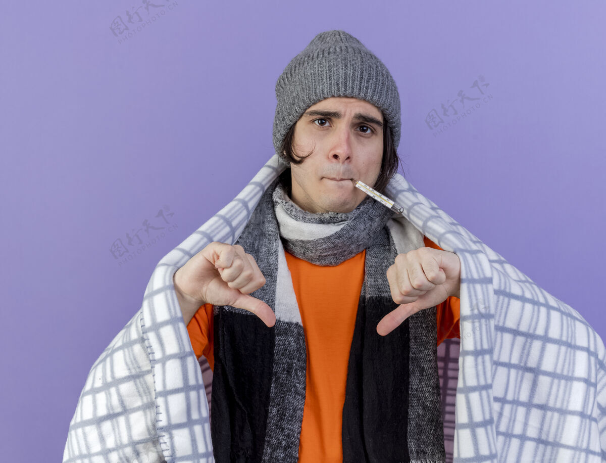 拇指未出狱的年轻病人戴着冬天的帽子 围巾裹在格子布里 嘴里叼着温度计 大拇指朝下 孤立地站在紫色地上男人紫色围巾