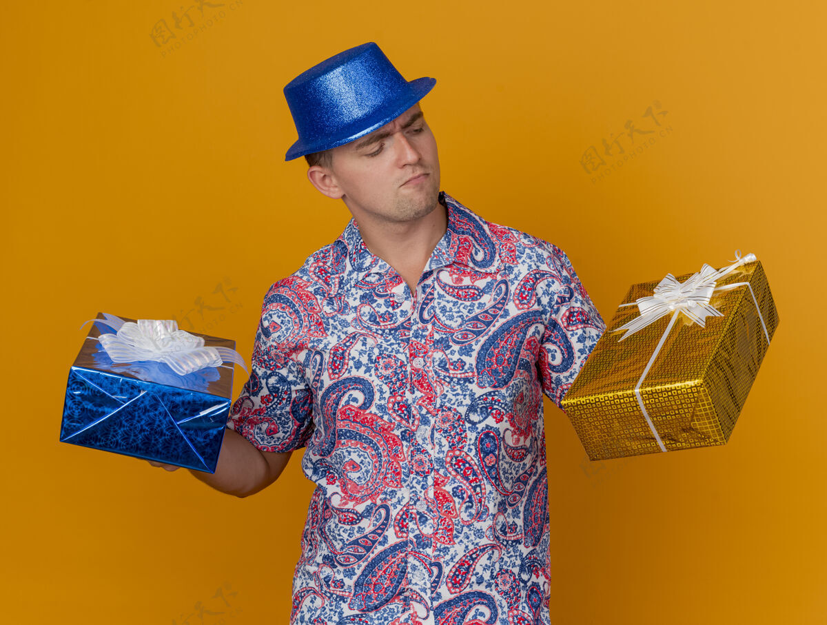 派对一个体贴的年轻人戴着蓝色的帽子 拿着礼物盒 看着孤立的橙色年轻盒子戴着