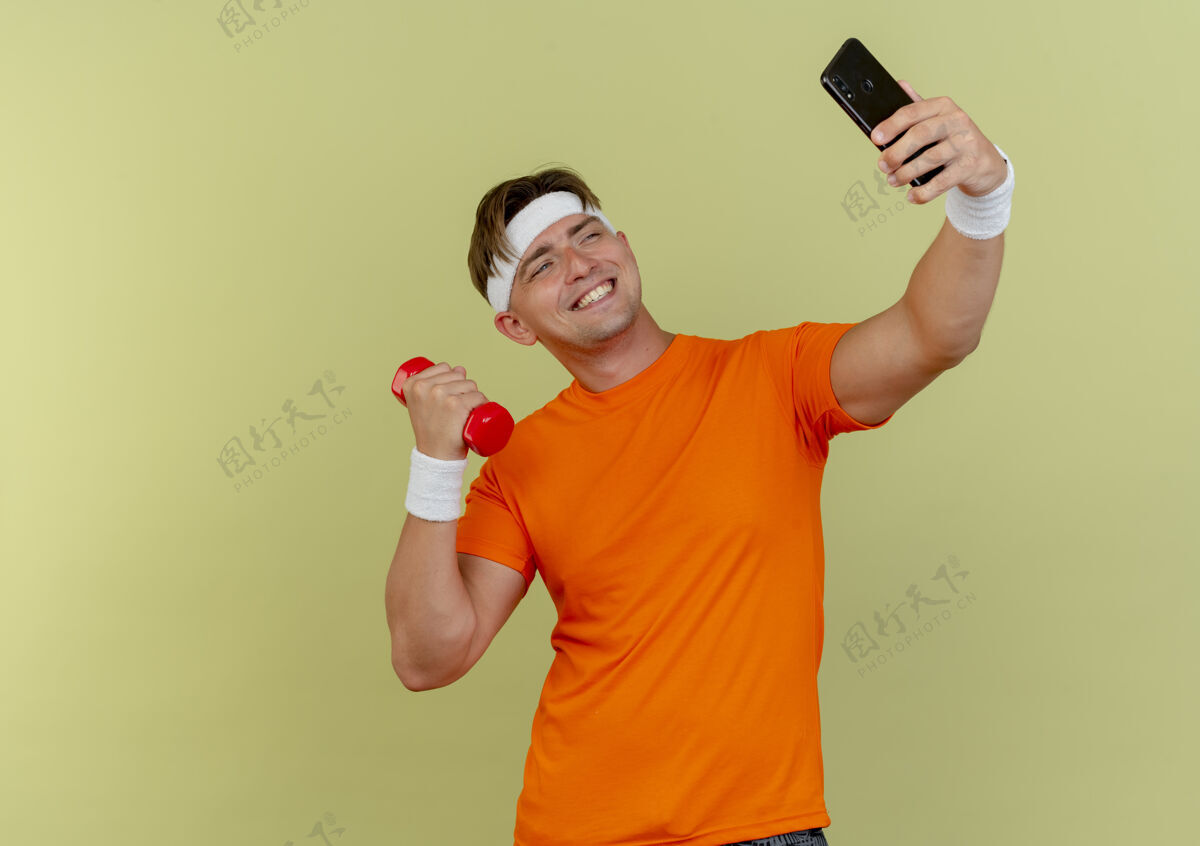 人微笑的年轻帅气的运动型男人戴着头带和腕带拿着手机和哑铃在橄榄绿的墙上自拍拿微笑绿色