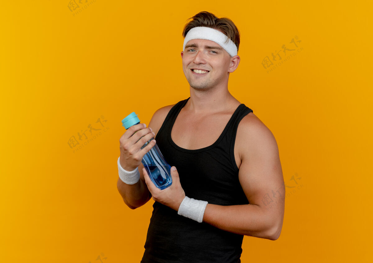拥抱面带微笑的年轻帅气的运动型男人戴着头带和手腕拿着水瓶隔离在橙色的墙上人人微笑