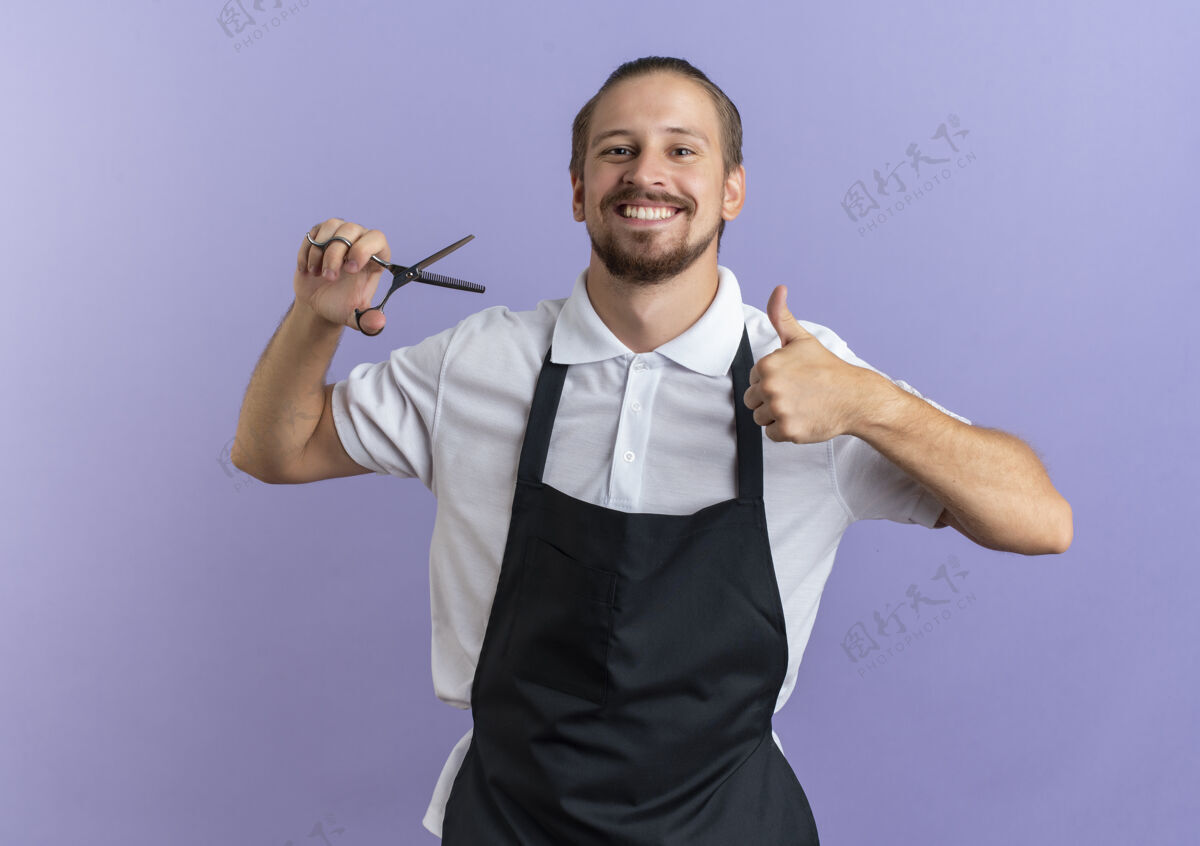 姿势面带微笑的年轻英俊的理发师穿着制服拿着剪刀 在紫色的墙上孤立地竖起大拇指紫色衣服帅气