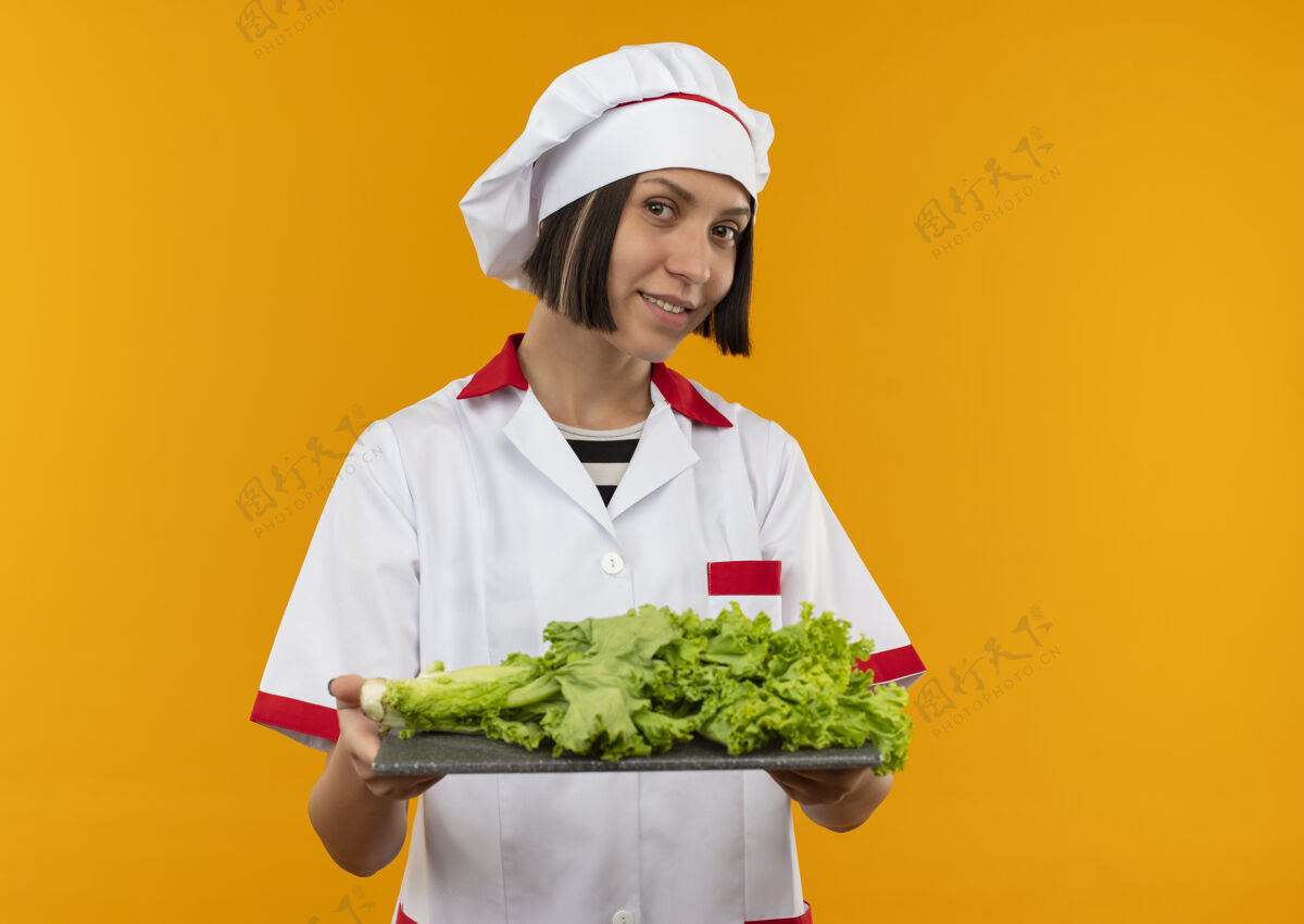 烹饪身着厨师制服 面带微笑的年轻女厨师手持砧板 砧板上有莴苣 隔离在橙色的墙上板姿势年轻