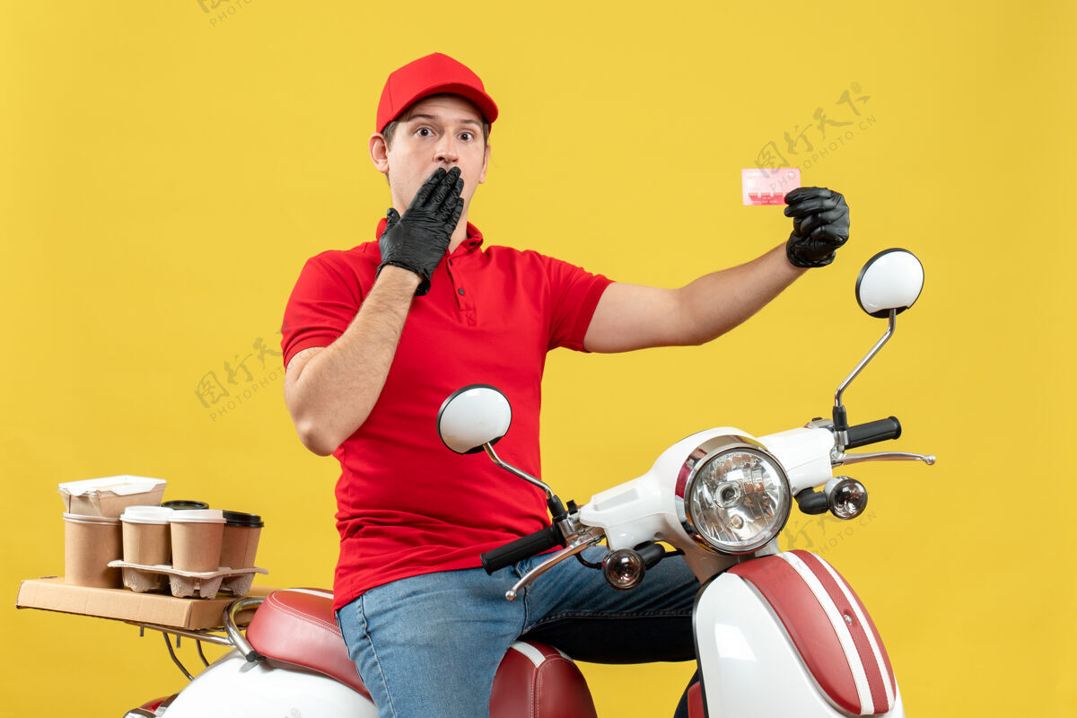 举行一个穿着红色上衣 戴着帽子手套 戴着医用面罩 坐在摩托车上拿着银行卡的快递员的正视图面罩命令人