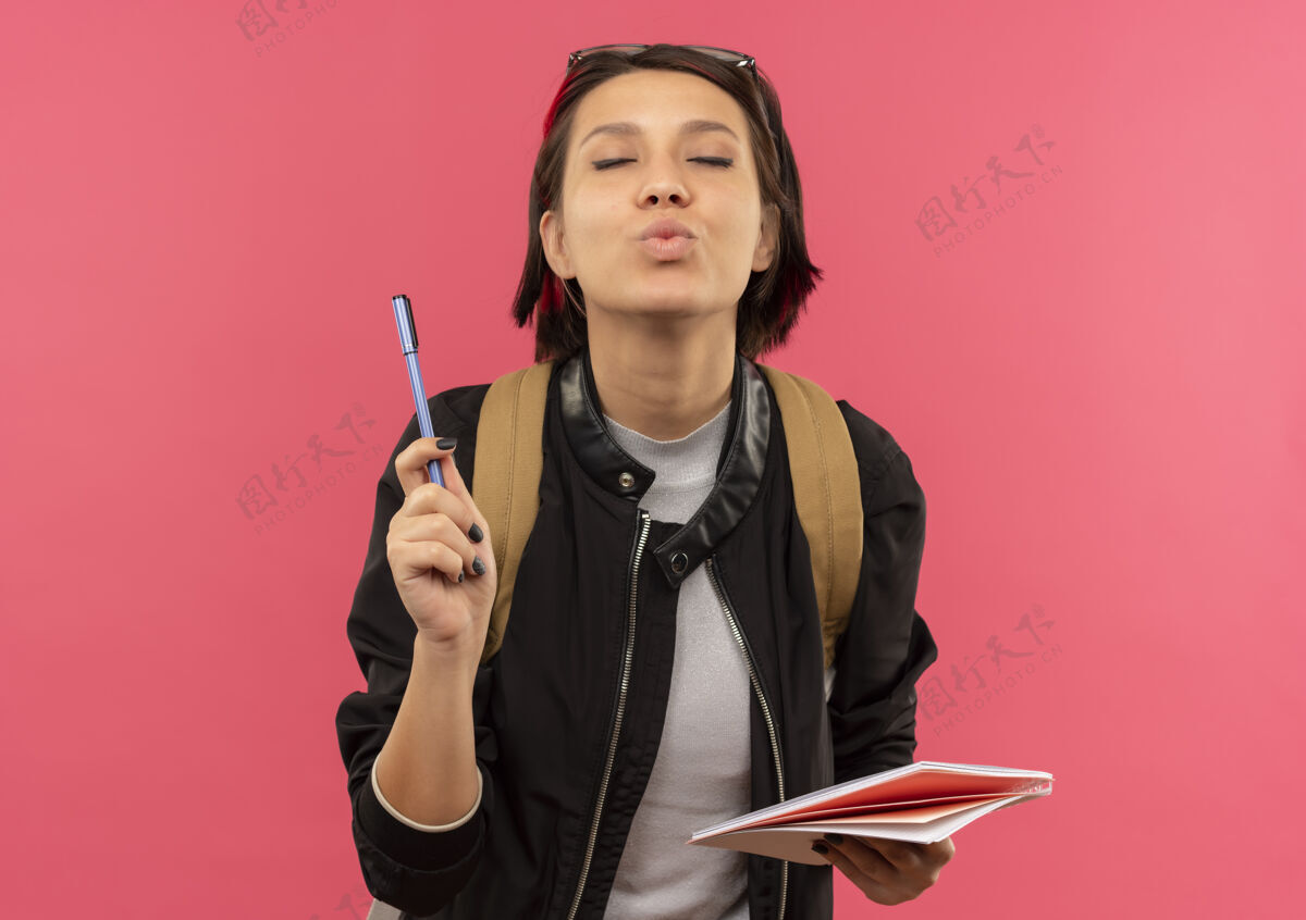 笔高兴的年轻女学生头戴眼镜 背着书包 手里拿着笔和记事本 闭着眼睛在粉红色的墙上亲吻粉色请衣服