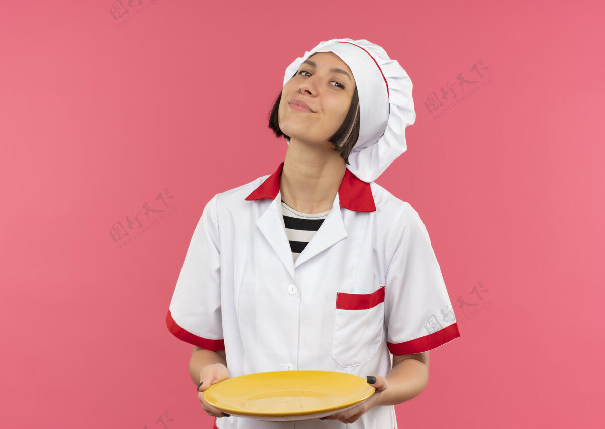 持有高兴的年轻女厨师在厨师制服举行空盘子隔离在粉红色的墙上空烹饪感情