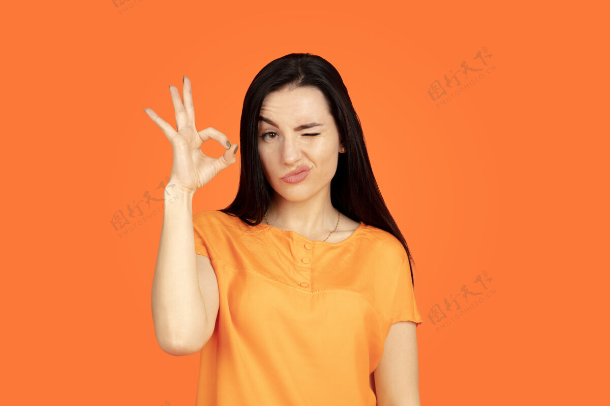 成功橙色工作室背景上的白种人年轻女子肖像穿着衬衫的漂亮深色女性模特人类情感的概念 面部表情 销售 广告广告空间显示ok的标志人类商务女性面部