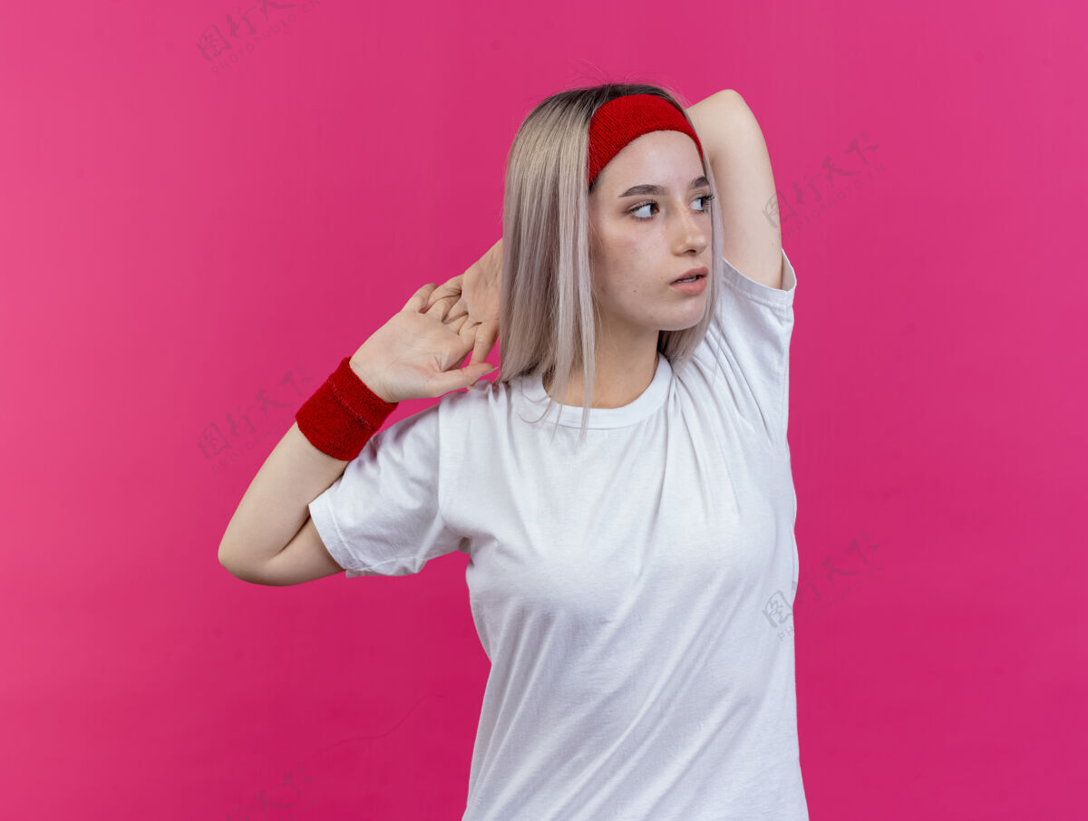 脖子自信的年轻运动女性戴着背带 戴着头带和腕带 双手合十放在脖子后面 看着粉色墙壁上孤立的一面穿女人运动