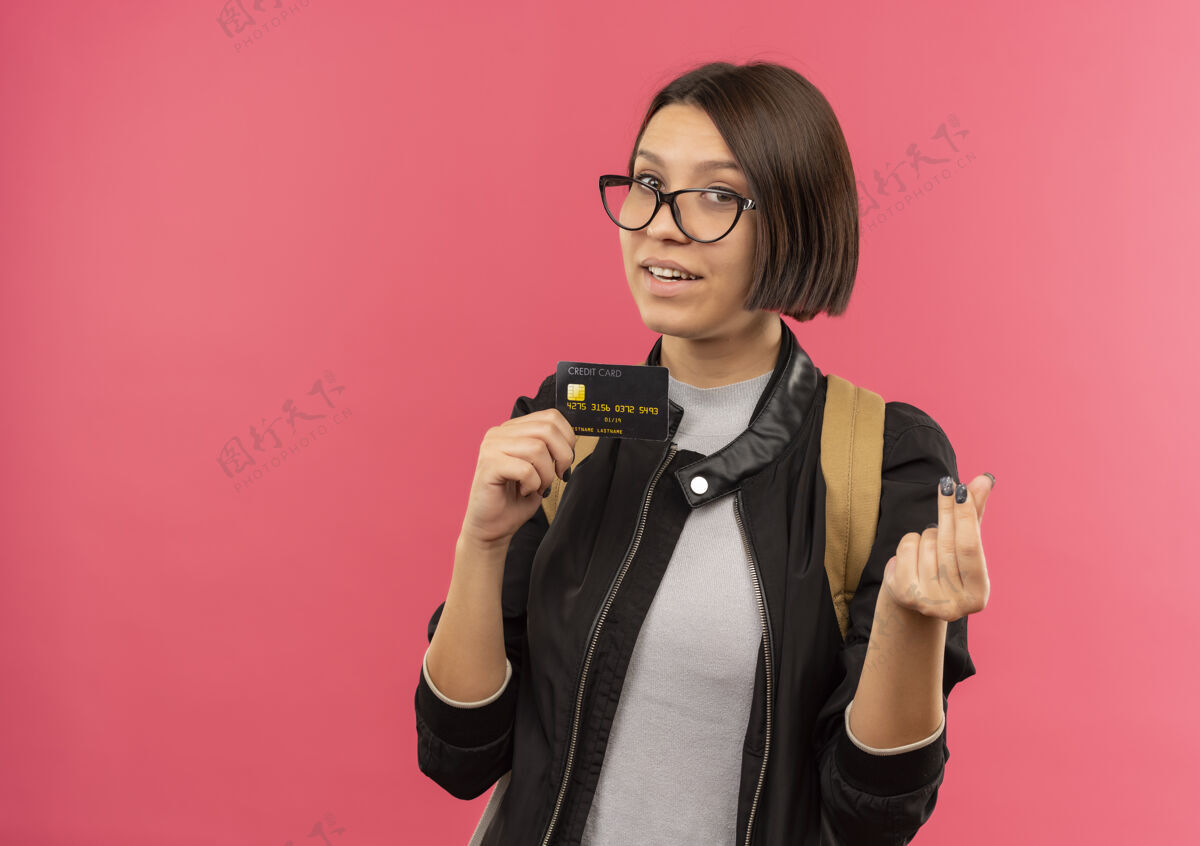 人高兴的年轻学生女孩戴着眼镜 背着包拿着信用卡 指着隔离在粉红色墙上的钱手势粉色包