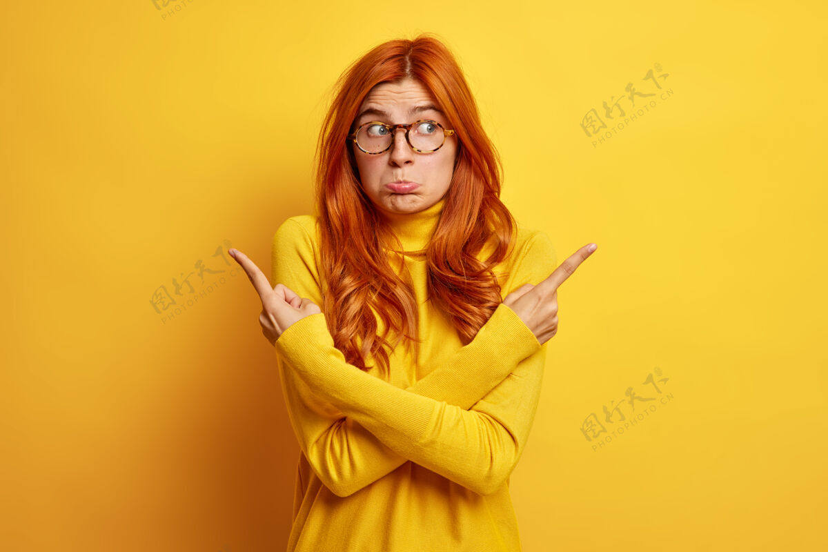右困惑不快乐的女人 长着姜黄色的头发 双臂交叉在身上 指向不同的侧面 犹豫着最好选择穿黄色的套头衫壁板手势黄色