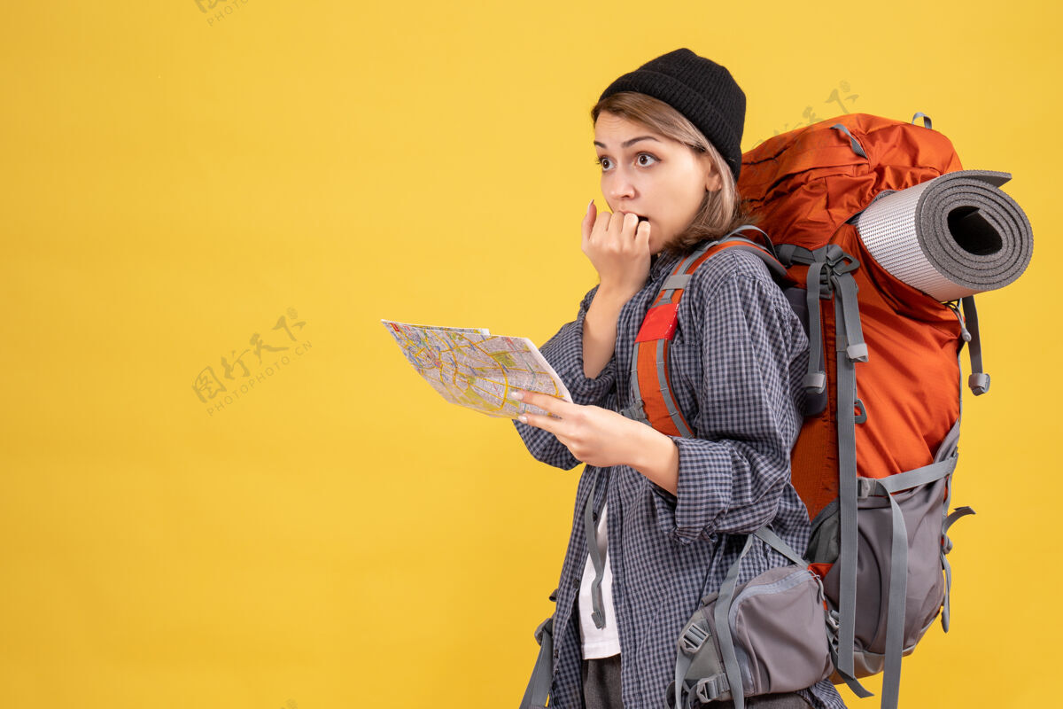 快乐困惑的年轻旅行者背着背包 拿着地图站在黄色的墙上微笑成人背包