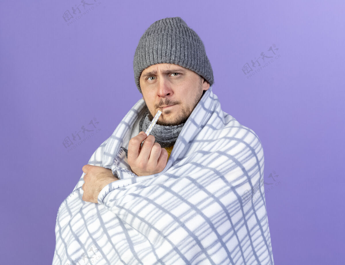 格子无知的年轻金发病斯拉夫男子冬季帽子和围巾裹在格子测量温度与温度计隔离在紫色墙上复制空间金发男人围巾