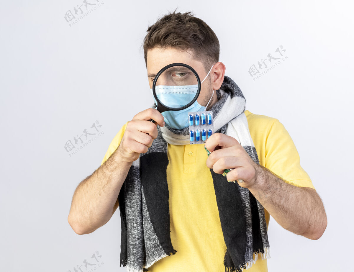 金发自信的年轻金发病斯拉夫男子戴着医用口罩和围巾看着通过放大镜在白色墙上的复制空间隔离药片包药片男人斯拉夫