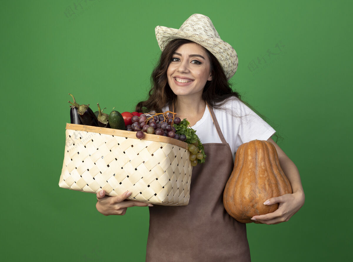 制服微笑着的年轻女园丁穿着制服 戴着园艺帽 手里拿着蔬菜篮子和南瓜 隔离在绿色的墙上 留着复制空间年轻南瓜花园