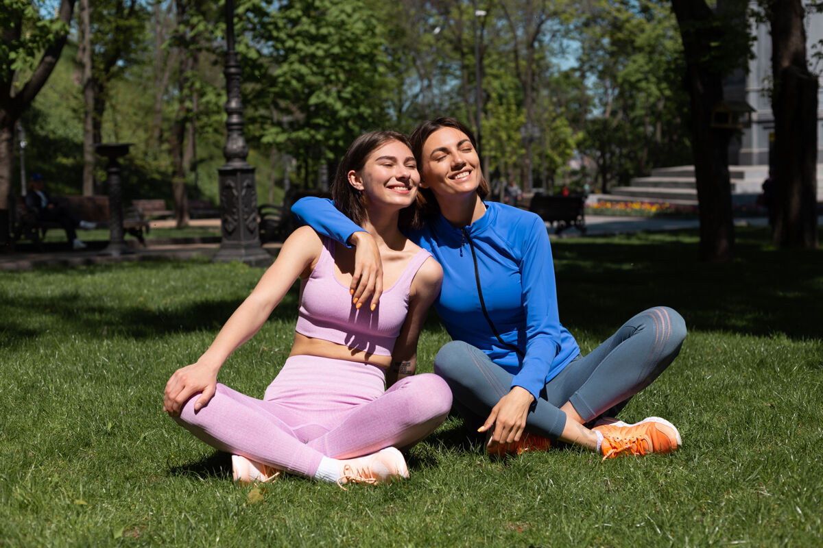 女性阳光明媚的日子里 两个穿着运动服的漂亮女人在公园的草地上做瑜伽 脸上带着微笑拥抱着对方活跃朋友健康