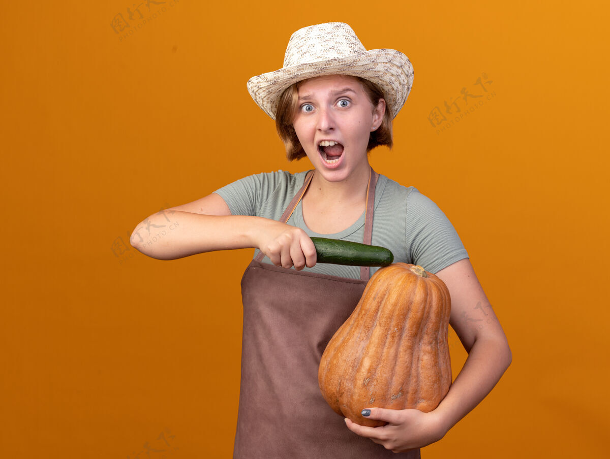 斯拉夫焦急的年轻的斯拉夫女园丁戴着园艺帽 手里拿着黄瓜和橘子上的南瓜南瓜帽子黄瓜