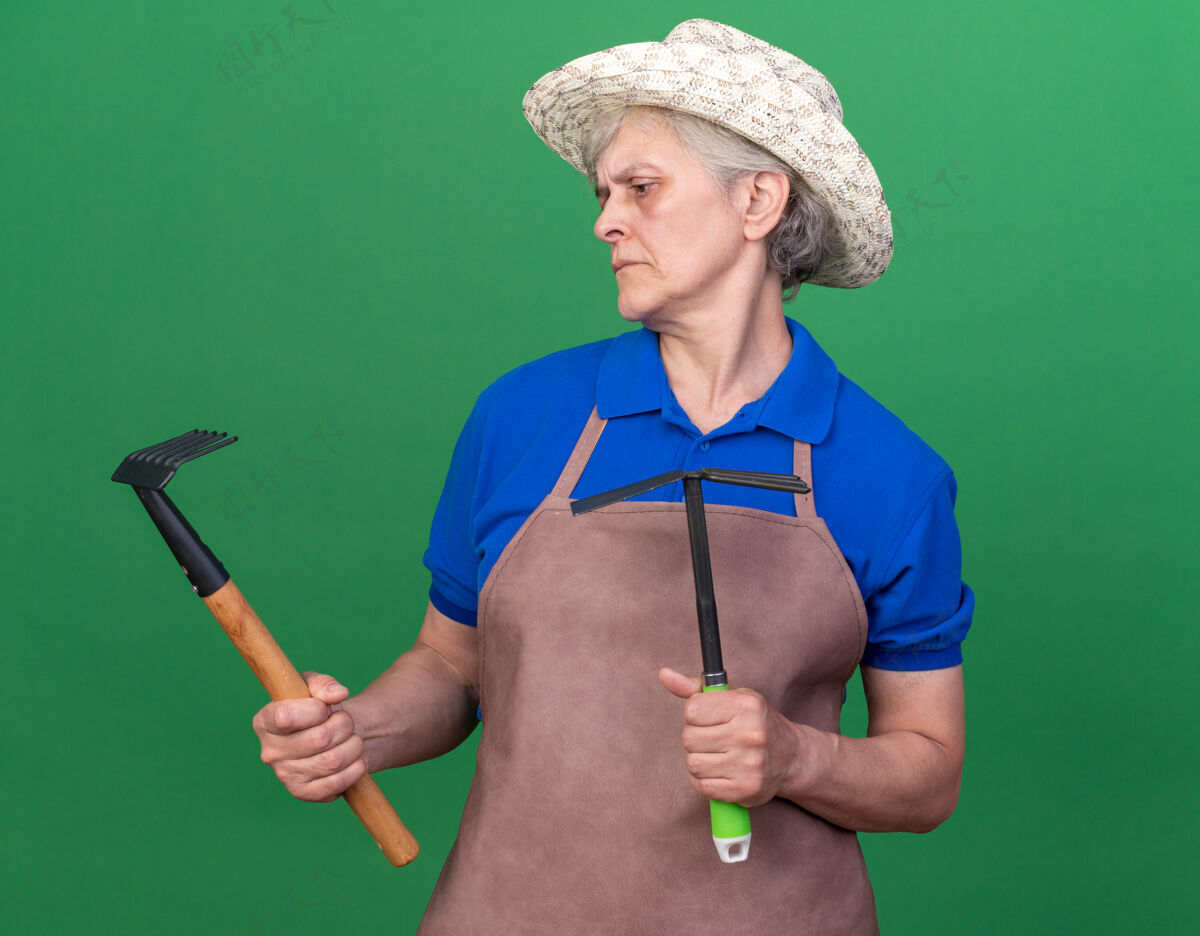 无知笨手笨脚的上了年纪的女园丁戴着园艺帽拿着锄头耙子看着耙子上的绿色园艺帽子老人