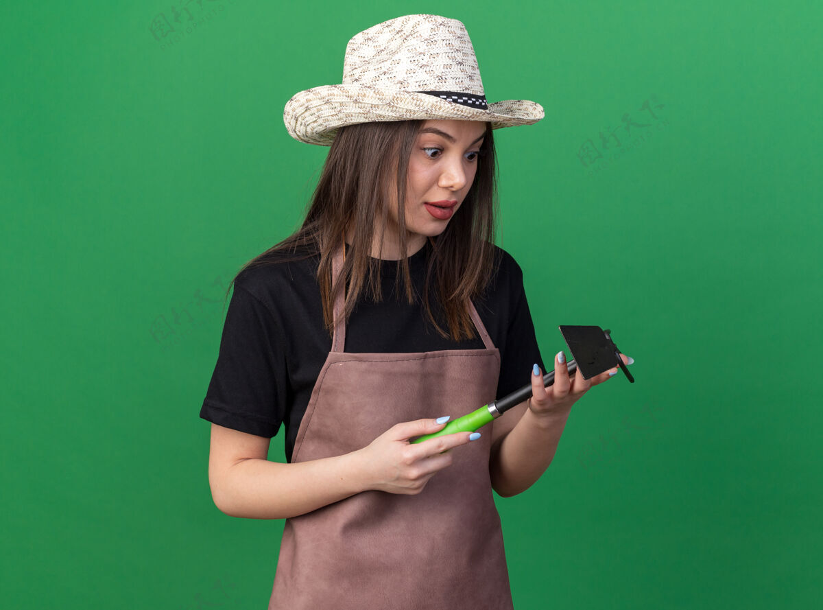 女漂亮的白人女园丁戴着园艺帽 手持锄头 看着耙子上的绿色帽子耙子园艺