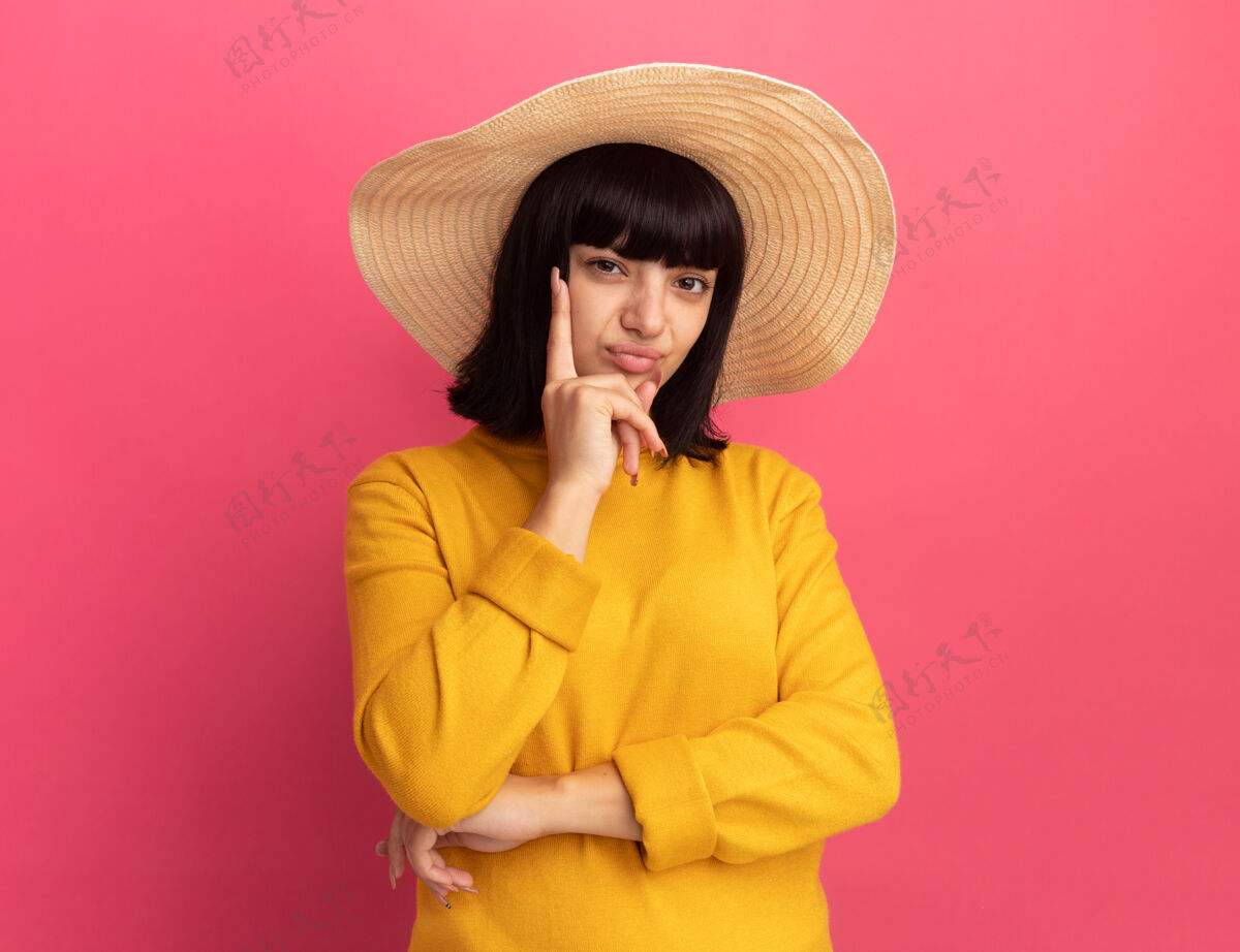 黑发年轻的黑发白人女孩戴着沙滩帽 手放在下巴上 看着粉红色的相机海滩令人讨厌帽子