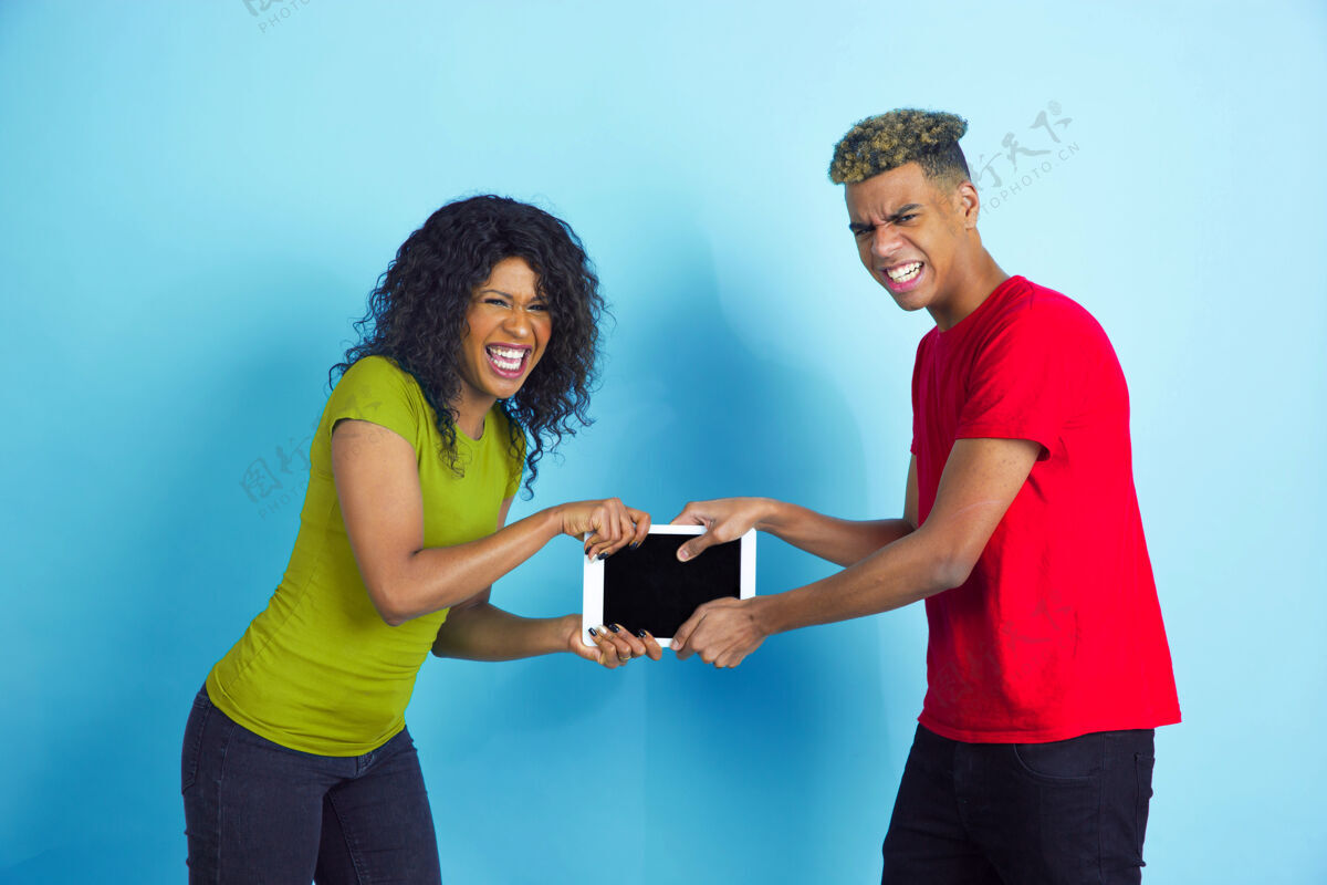 面部为好玩的平板电脑而战年轻情绪化的非裔美国人男女穿着蓝色背景的彩色休闲服美丽的情侣人类情感的概念 面部表情 关系 广告朋友头发衣服