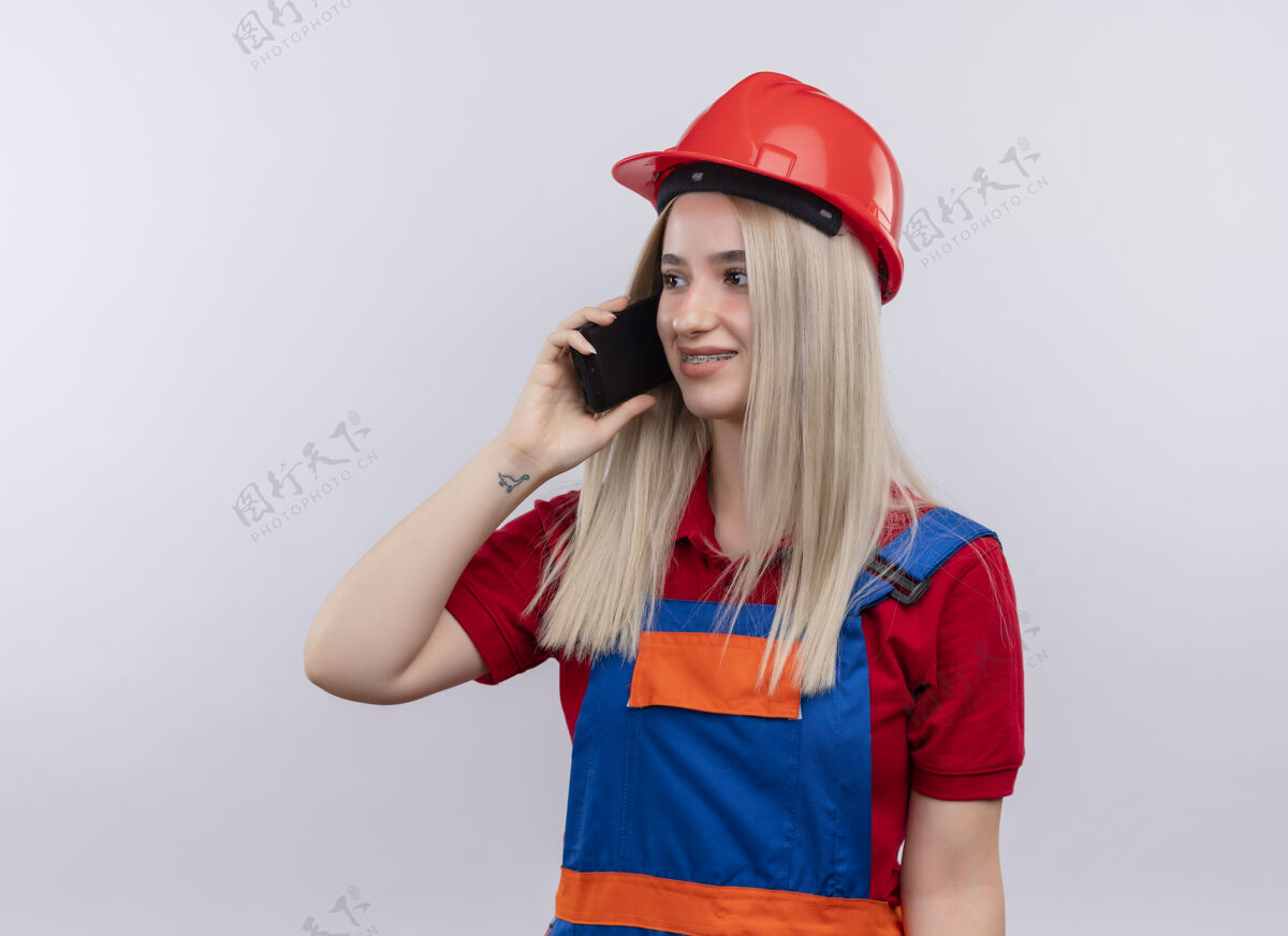 工程师微笑着的年轻金发工程师建筑工女孩穿着制服戴着牙套在电话里说话看着左边的空白处和拷贝空间微笑年轻建筑工人