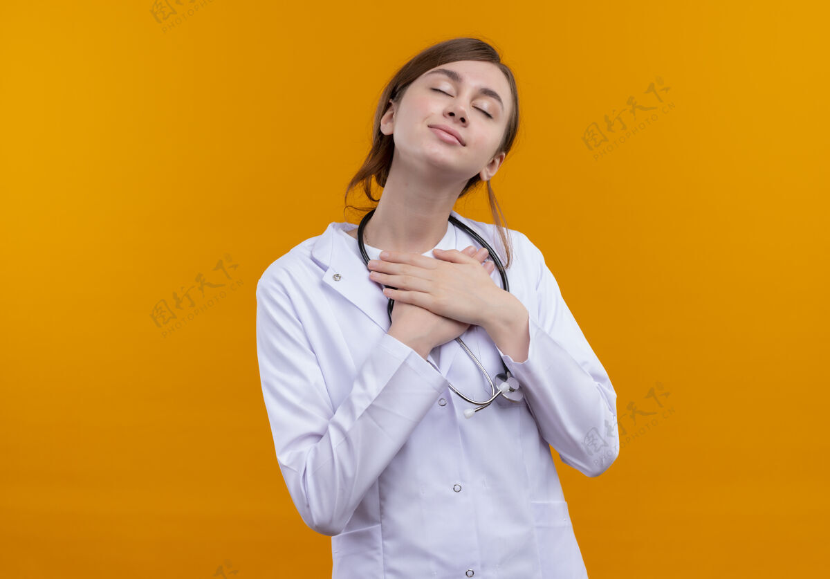 手穿着医用长袍和听诊器的年轻女医生 双手放在胸前 闭上眼睛 在孤立的橙色空间里 有复印空间副本医生和平