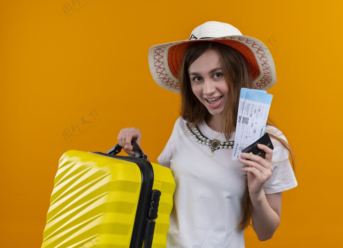 橙色快乐的年轻旅客女孩戴着帽子 拿着手提箱和机票 信用卡在孤立的橙色空间手提箱持有女孩