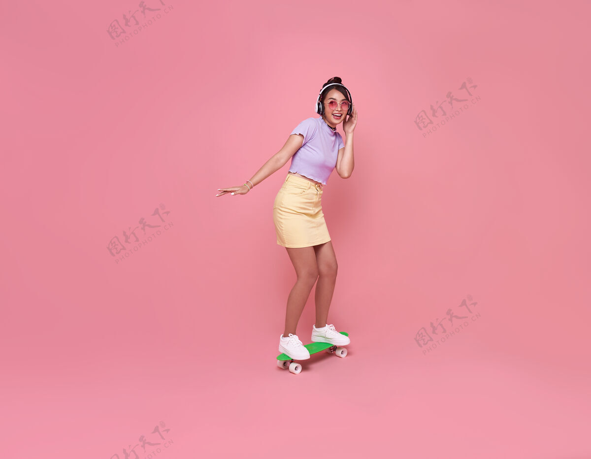 乐趣年轻的亚洲少女戴着无线耳机在粉红色的墙上玩滑板 听音乐时尚女人肖像