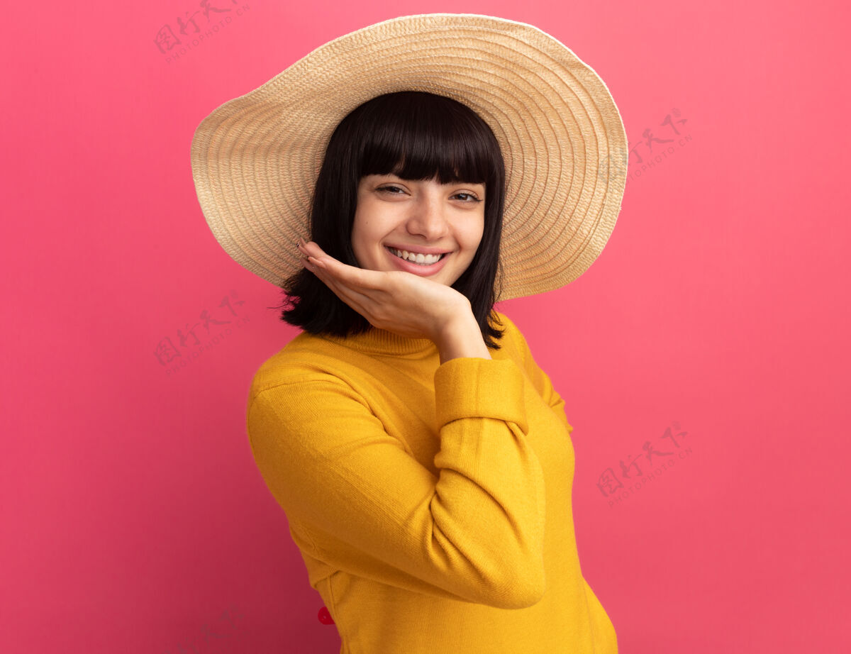 年轻年轻的黑发白人女孩戴着沙滩帽 高兴地把手放在下巴上 看着粉红色的相机沙滩拜托帽子