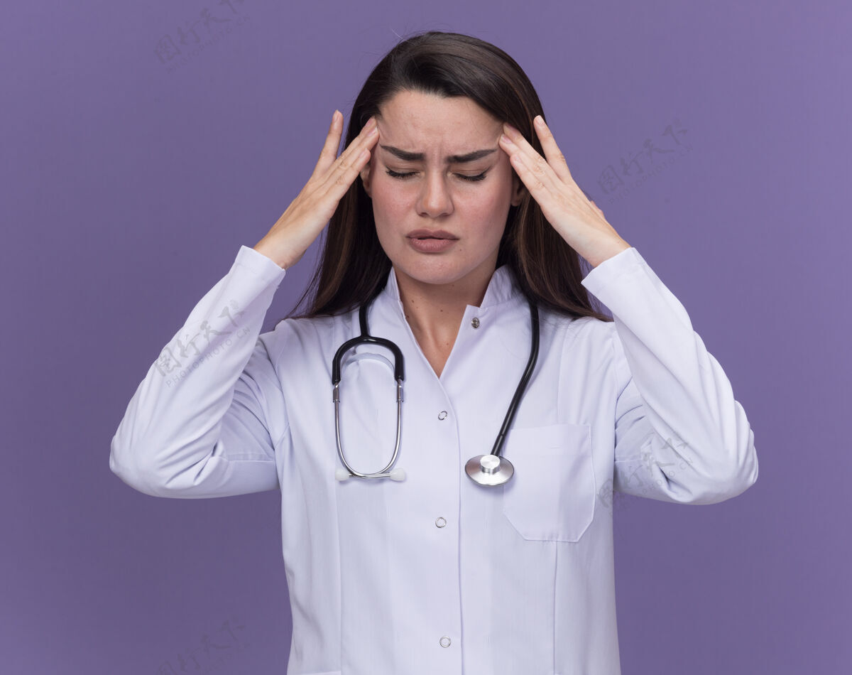 疼痛疼痛的年轻女医生穿着带听诊器的医用长袍 双手放在头上 闭着眼睛站在紫色上年轻女性医生