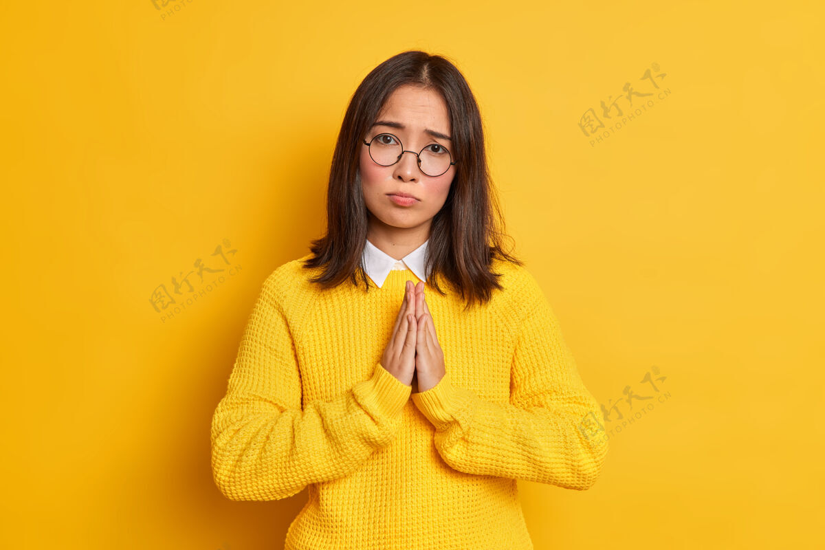 黑发乞讨的亚洲年轻女子 黑发 双手保持祈祷姿势 表情恳求 戴着圆眼镜 穿着休闲毛衣韩国人旗帜穿着