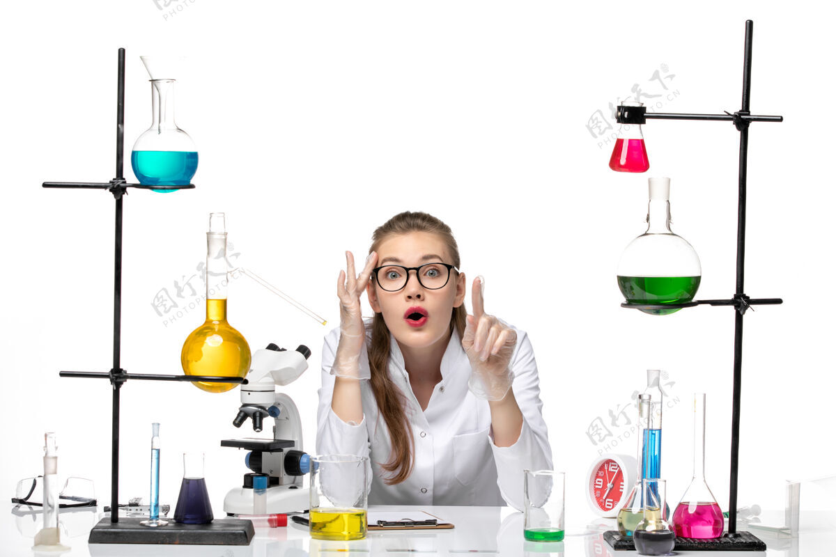 研究前视图：身着医疗服的女化学家坐在桌子前 用浅白背景上的溶液化学大流行性冠状病毒实验白色医学
