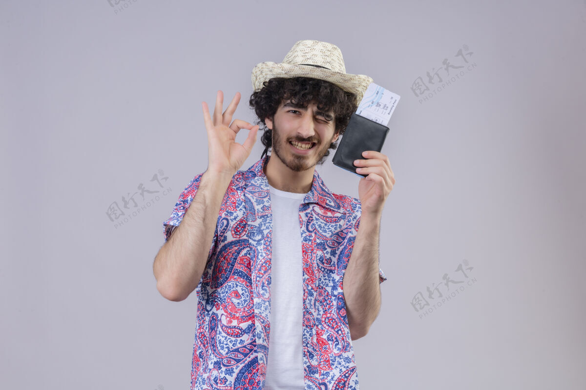 旅行者自信的年轻帅气的卷发旅行者戴着帽子做着“ok”的手势 在隔离的空白处展示钱包和机票展示标志机票