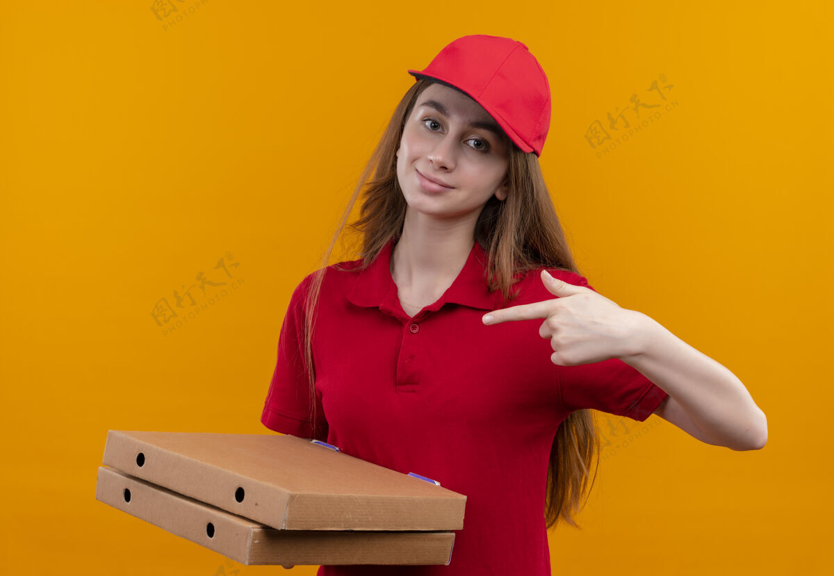 女孩在孤立的橙色空间里 穿着红色制服的自信的年轻送货女孩拿着包裹指着他们盒子橙色拿着