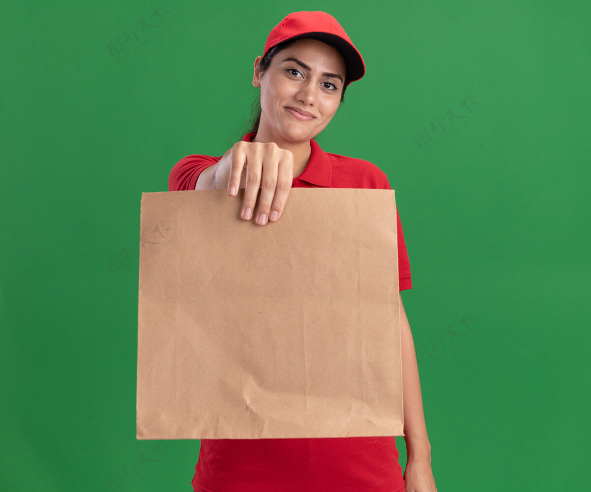 脸高兴的年轻送货女孩穿着制服 戴着帽子 手里拿着纸食品包 在绿色的墙前孤立着食物站着人