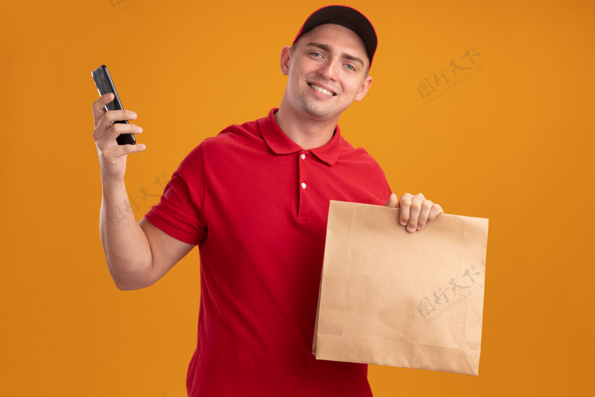 衣服带着微笑的年轻送货员 穿着制服 戴着帽子 拿着纸食品包 电话被隔离在橙色的墙上递送年轻人纸
