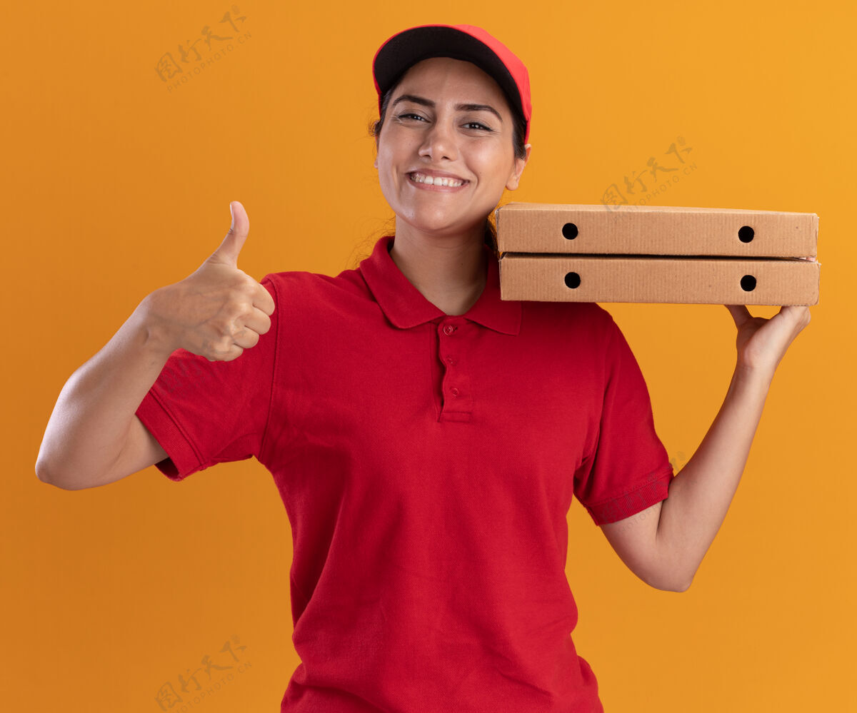 人微笑着的年轻送货女孩穿着制服 戴着帽子 肩上扛着披萨盒 在橙色的墙上孤立地竖起大拇指女孩公民脸