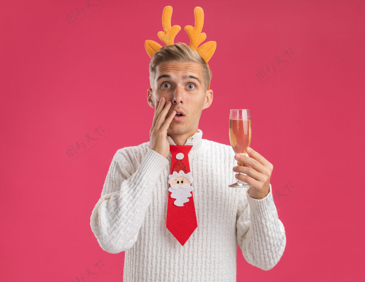 触摸关心的年轻帅哥戴着驯鹿鹿角头带打着圣诞老人的领带拿着一杯香槟看着镜头抚摸着隔离在粉色背景上的脸粉色鹿角圣诞快乐