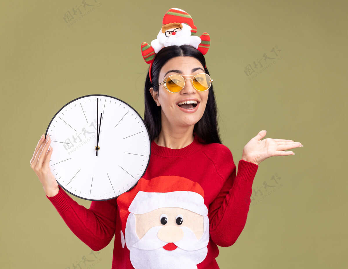 绿色快乐的年轻白种女孩戴着圣诞老人的头带和毛衣 戴着眼镜 拿着时钟 看着相机 在橄榄绿的背景上孤立着一只空手时钟头带毛衣