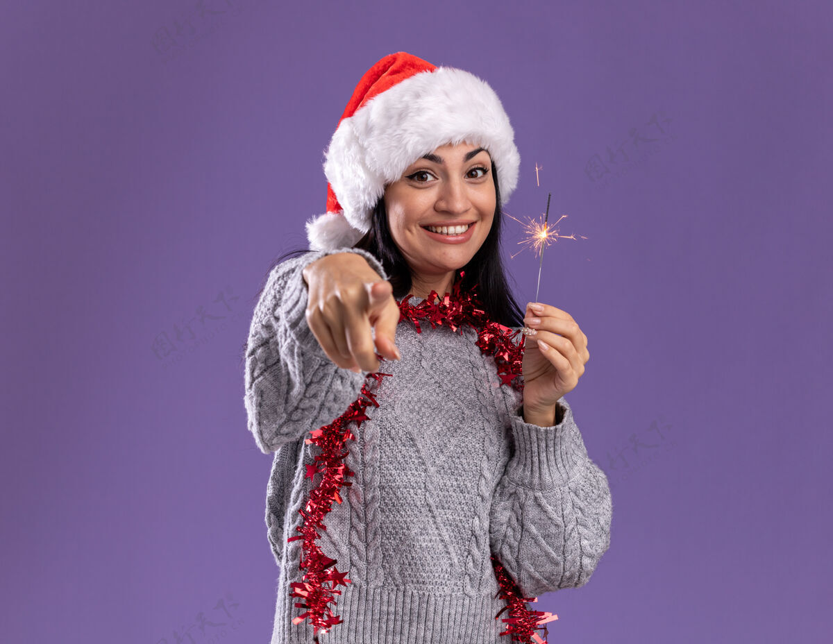 新年快乐的年轻白人女孩戴着圣诞帽 脖子上戴着金箔花环 手里拿着节日火花 看着和紫色背景隔离的相机年轻圣诞节帽子