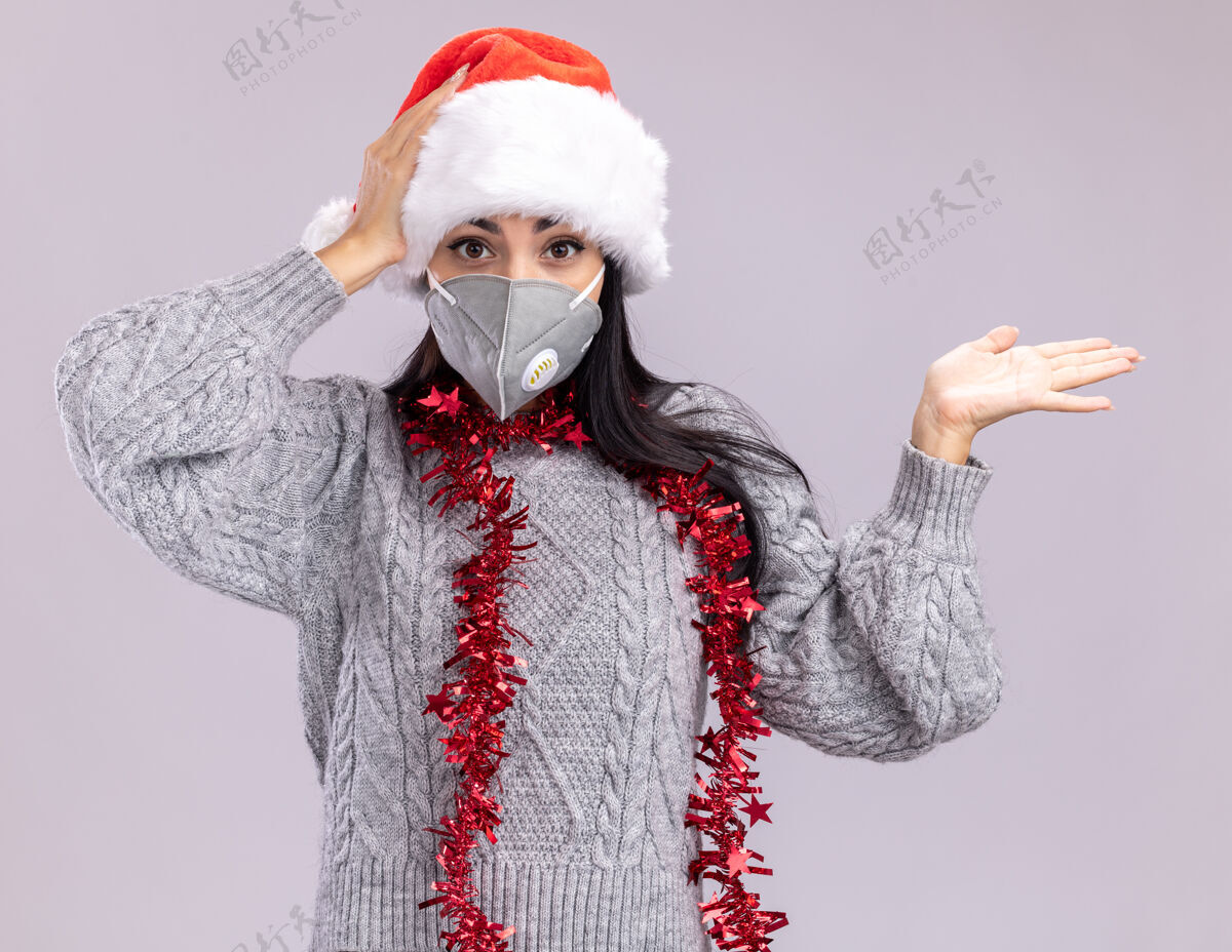 保护关心的年轻白人女孩戴着圣诞帽 脖子上戴着金属丝花环 戴着防护面具 看着镜头 一只手空着 另一只手放在头上 与白色背景隔离镀金圣诞节圣诞快乐