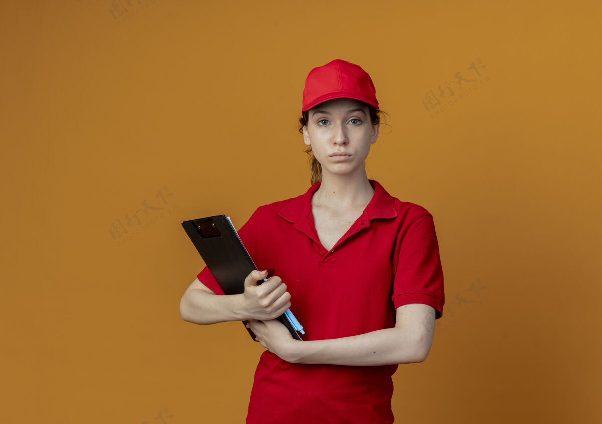 女孩年轻漂亮的送货女孩穿着红色制服 戴着帽子 看着相机拿着剪贴板和笔 孤立地放在橙色背景上 还有复印空间红色制服剪贴板