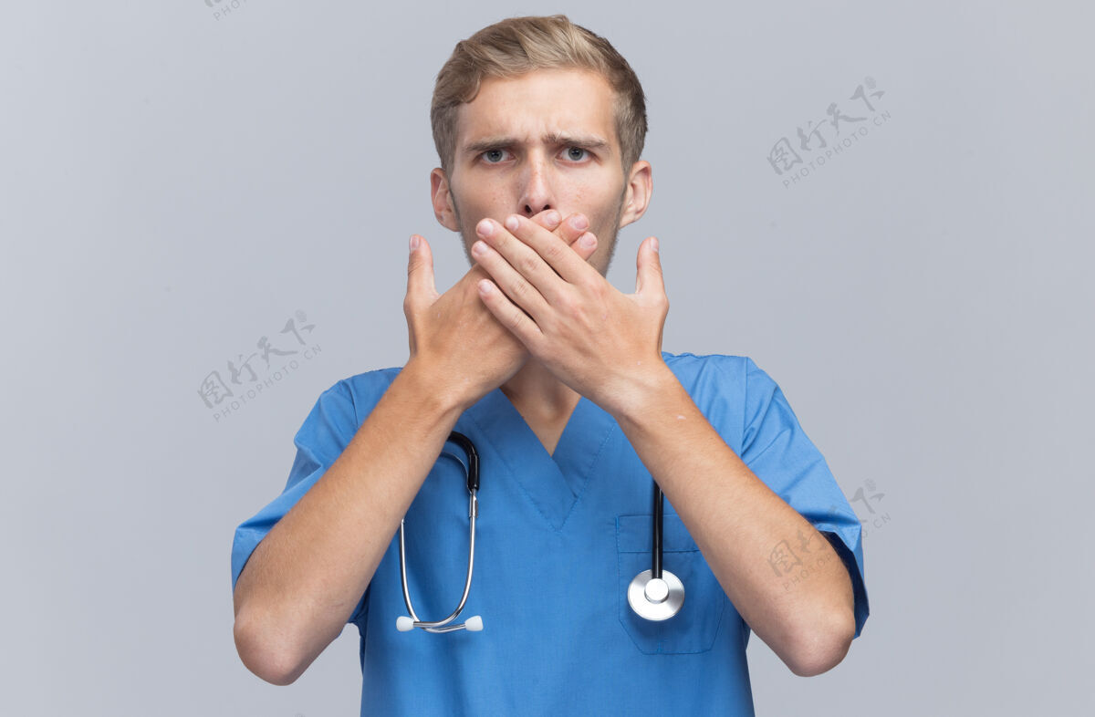 人害怕的年轻男医生穿着医生制服用听诊器捂住嘴双手隔离在白墙上掩护站着嘴