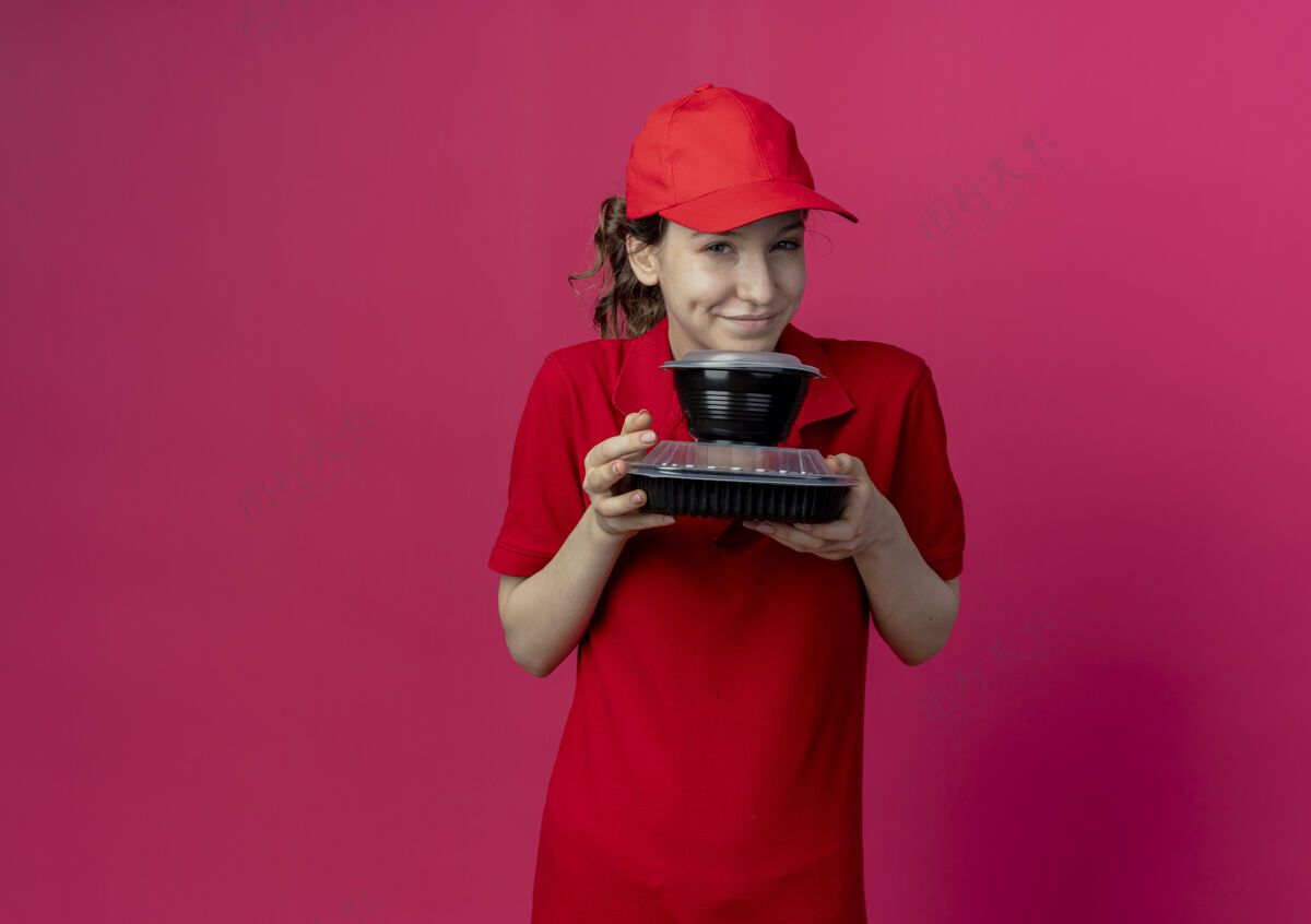 请令人高兴的年轻漂亮的送货女孩穿着红色制服 戴着帽子 拿着食物容器 在深红色的背景上与复制空间隔离背景红色制服